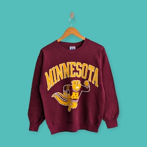 May include: A maroon sweatshirt with the word "MINNESOTA" in gold arched lettering. Below the text is a cartoon gopher mascot in a gold and maroon uniform. The sweatshirt is hanging on a wooden hanger against a teal background.