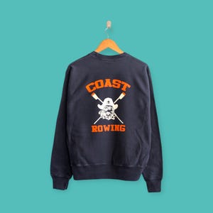 Puede incluir: Sudadera azul marino con las palabras "COAST ROWING" en letras naranjas. El diseño presenta una calavera pirata con remos cruzados. La sudadera está colgada en una percha de madera sobre un fondo turquesa.