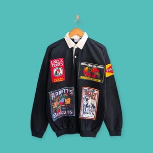 Pode incluir: Camisa preta de manga comprida com gola e vários patches de estilo vintage. Os patches incluem texto e gráficos para "Uncle Tobys Oats", "Australasian Jam Company", "Arnott's Biscuits" e "Billy Tea". A camisa tem uma gola branca.