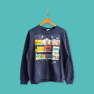 Puede incluir: Sudadera azul marino con un gráfico colorido de edificios de estilo suroeste. El gráfico presenta edificios con fachadas amarillas, naranjas, azules y verdes. La sudadera está colgada en una percha de madera sobre un fondo turquesa.