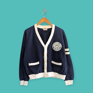 May include: Navy blue cardigan with white trim, pockets, and button closure. Features a patch on the chest with the text "UNIVERSITY OHIO IVY CLUB". The sleeves have white stripes. The cardigan is hanging on a wooden hanger.