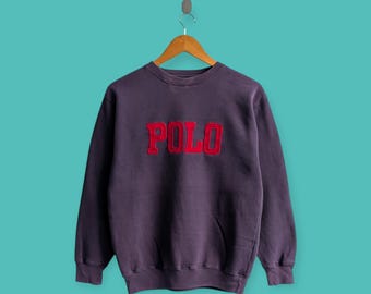 May include: A dark purple sweatshirt with the word "POLO" in red, block letters across the chest. The sweatshirt is hanging on a wooden hanger against a teal background. The garment has a vintage, worn appearance.
