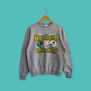 Puede incluir: Una sudadera gris jaspeado con un gráfico que conmemora el Campeonato Nacional Toshiba BCS 2011. El diseño presenta cascos de fútbol y el texto "NATIONAL CHAMPIONSHIP" en amarillo y verde. La sudadera tiene mangas largas.