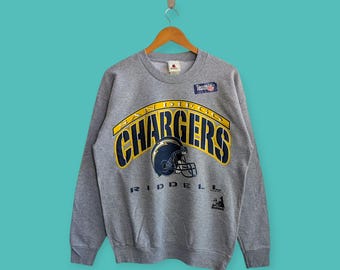 Puede incluir: Sudadera gris de cuello redondo con el logotipo de los San Diego Chargers en amarillo y azul marino. El diseño incluye el nombre del equipo y un casco de fútbol. La sudadera es de Riddell y tiene un aspecto vintage.