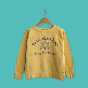 May include: Mustard yellow sweatshirt with the text "United States Army Army Air Forces" in black. The sweatshirt is hanging on a wooden hanger against a teal background. The design includes an eagle emblem.