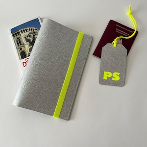 May include: A gray passport holder with a bright yellow elastic band. A matching gray luggage tag with the initials "PS" in yellow is also shown. The luggage tag is attached to a red passport with the text "Europ&auml;ische Union Bundesrepublik Deutschland" in white.