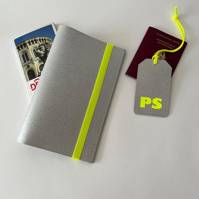 May include: A gray passport holder with a bright yellow elastic band. A matching gray luggage tag with the initials "PS" in yellow is also shown. The luggage tag is attached to a red passport with the text "Europ&auml;ische Union Bundesrepublik Deutschland" in white.