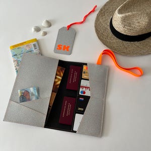 May include: A gray passport holder with a black interior and multiple pockets for cards, cash, and documents. The holder has a bright orange strap and a matching tag with the letters "SK" in orange.