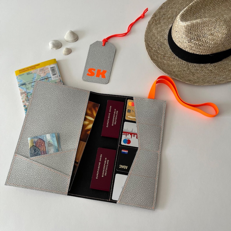 May include: A gray passport holder with a black interior and multiple pockets for cards, cash, and documents. The holder has a bright orange strap and a matching tag with the letters "SK" in orange.