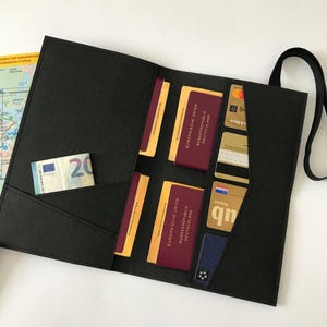 May include: Black travel wallet with multiple pockets holding passports, credit cards, and a twenty-euro bill. The wallet has a black strap and is open, displaying the contents. A map is visible in the background.