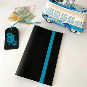 May include: Black notebook with a turquoise elastic band. A black tag with turquoise floral design hangs nearby. A blue and white bus-shaped piggy bank with a surfboard on top is in the background. Currency and coins are also visible.