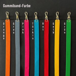 May include: A selection of colorful elastic bands, each clipped with a gold-colored clip. The bands are orange, gray, pink, turquoise, red, yellow, and green. The German text "Gummiband-Farbe" is at the top.