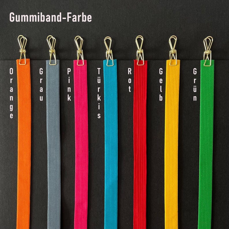 May include: A selection of colorful elastic bands, each clipped with a gold-colored clip. The bands are orange, gray, pink, turquoise, red, yellow, and green. The German text "Gummiband-Farbe" is at the top.