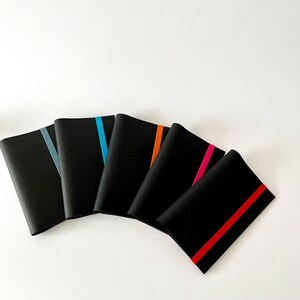 May include: A collection of five black notebooks with colorful accent stripes. Each notebook has a different stripe color: gray, blue, orange, pink, and red. The notebooks are arranged in a fan shape against a white background.