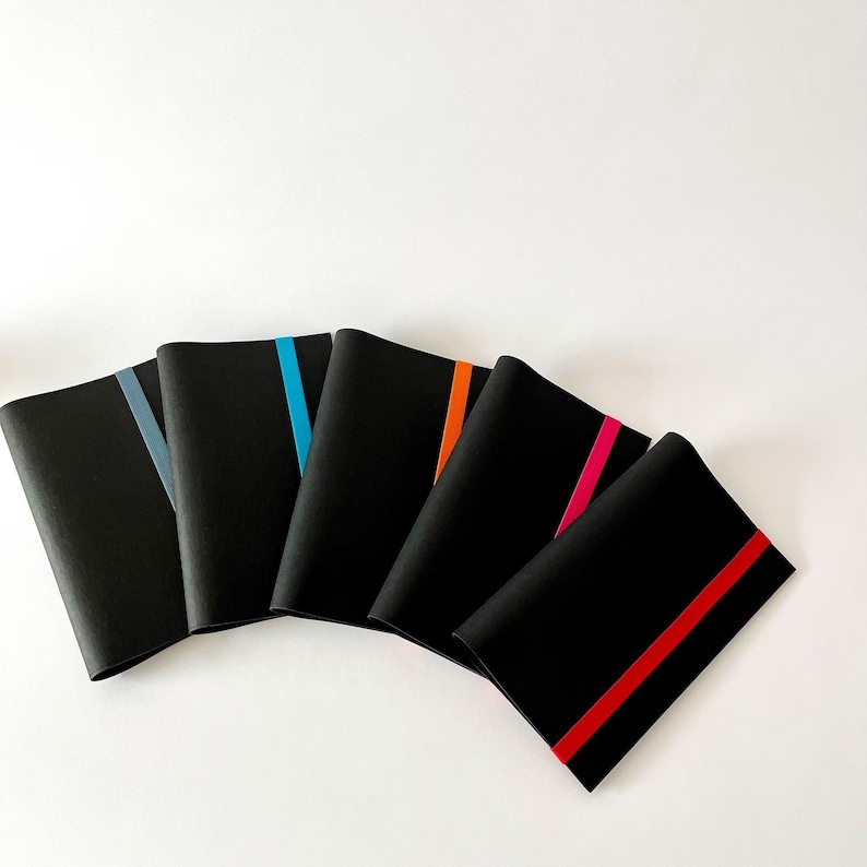 May include: A collection of five black notebooks with colorful accent stripes. Each notebook has a different stripe color: gray, blue, orange, pink, and red. The notebooks are arranged in a fan shape against a white background.