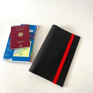 May include: A black passport holder with a red elastic band, next to a stack of travel guides and a burgundy passport. The passport is labeled "REISEPASS" and has a gold emblem.