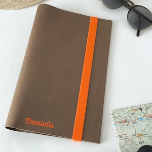 Könnte beinhalten: Ein braunes Notizbuch mit einem vertikalen orangefarbenen Gummibandverschluss. Der Name Daniela ist in Orange in der unteren linken Ecke des Einbands aufgedruckt. Das Notizbuch liegt neben einer Sonnenbrille mit dunklem Rahmen und einer Karte.