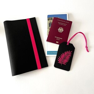 May include: A black passport cover with a bright pink elastic band, a burgundy passport, and a black luggage tag with a pink leaf design. The passport is open to a page with text. A white card with text is also visible.