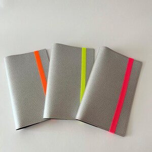 May include: Three gray leather passport holders with a contrasting neon colored stripe. The stripes are orange, yellow, and pink.