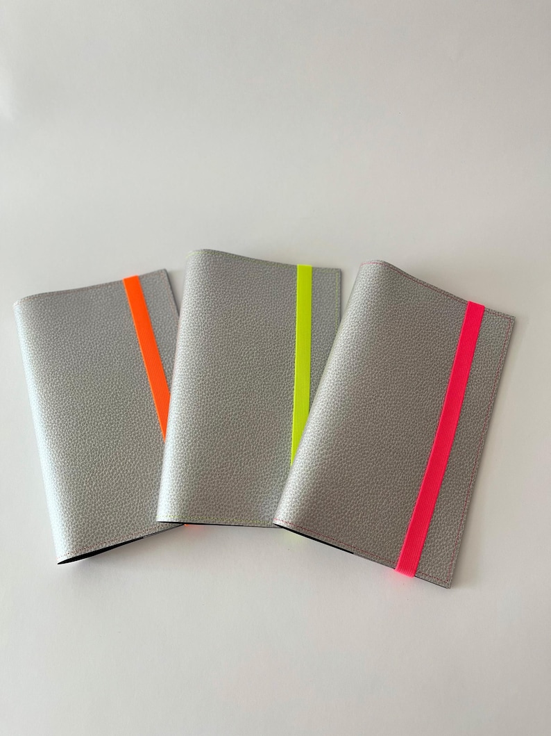 May include: Three gray leather passport holders with a contrasting neon colored stripe. The stripes are orange, yellow, and pink.