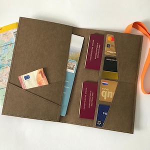 Könnte beinhalten: Ein braunes Reise-Portemonnaie mit mehreren Kartenfächern, Passhaltern und einer Tasche mit einem Euro-Schein. Ein orangefarbener Riemen ist befestigt. Eine Karte ist links sichtbar.