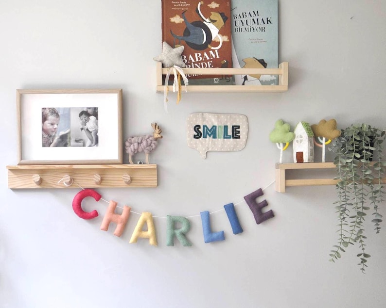 May include: A rainbow felt letter banner spelling "CHARLIE" hangs on a wall with wooden shelves. The shelves are decorated with a framed photo, a small stuffed animal, a felt word art piece that says "SMILE", and a small house with trees.