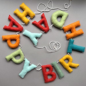 May include: A colorful felt banner with the word "Happy Birthday" spelled out in individual letters. The letters are strung together with white twine.