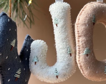 May include: Close-up of three felt Christmas ornaments spelling "JOY". The letters are in shades of blue, white, and tan, each decorated with embroidered Christmas trees. The ornaments hang from white ribbons, perfect for holiday decor.