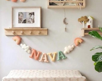 May include: A felt garland spelling the name NAVA in pastel letters, accented with clouds and rainbows, hanging below wooden wall shelves. The shelves hold books, a framed photograph, a small house figurine, and a banner of felt raindrops.