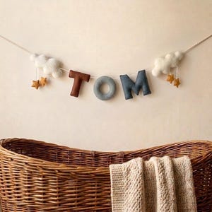Könnte beinhalten: Eine Filzbuchstaben-Girlande mit dem Namen TOM, bestehend aus einem braunen T, einem grauen O und einem dunkelgrauen M. Die Girlande ist an beiden Enden mit zwei weißen Wolken verziert, an denen jeweils zwei kleine senfgelbe Sterne hängen.