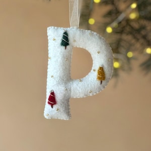 May include: A white felt letter P ornament featuring embroidered miniature trees in dark green, mustard yellow, and red. The letter is decorated with small gold beads and hangs from a thin, translucent ribbon loop for display purposes.