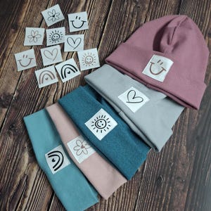 Könnte beinhalten: Vier verschiedene farbige Beanies mit verschiedenen gestickten Designs. Die Beanies sind rosa, grau, türkis und hellrosa. Die Designs beinhalten einen Regenbogen, eine Blume, eine Sonne und ein Herz.
