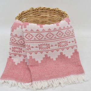 May include: A pink and white patterned Turkish towel with fringe. The towel is folded and draped over a wicker basket.