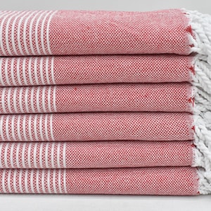 以下が含まれることがあります： 赤と白のストライプ柄のタオルが6枚重ねられています。タオルは織り込まれた質感で、端には白いフリンジが付いています。タオルはきちんと折りたたまれ、交互に配置された赤と白のストライプが際立っています。