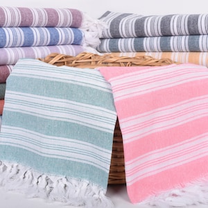 Puede incluir: Montones de toallas de playa a rayas en varios colores, incluyendo rosa, azul y gris. Las toallas tienen flecos blancos y est&aacute;n dispuestas en pilas ordenadas. Las toallas est&aacute;n hechas de un material suave.