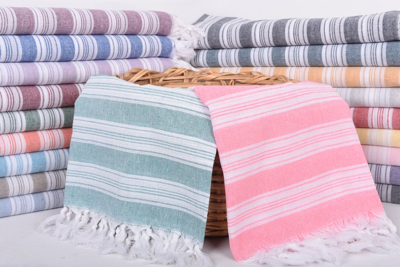 Puede incluir: Montones de toallas de playa a rayas en varios colores, incluyendo rosa, azul y gris. Las toallas tienen flecos blancos y est&aacute;n dispuestas en pilas ordenadas. Las toallas est&aacute;n hechas de un material suave.