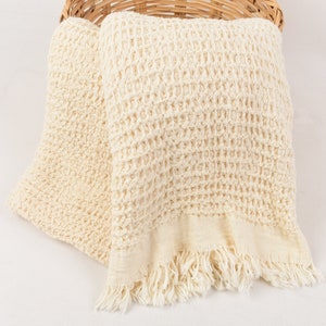 May include: A cream-colored waffle weave throw blanket with fringe edges. The blanket is folded and placed in a wicker basket.