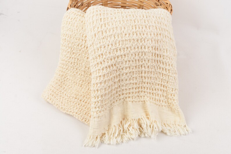 May include: A cream-colored waffle weave throw blanket with fringe edges. The blanket is folded and placed in a wicker basket.