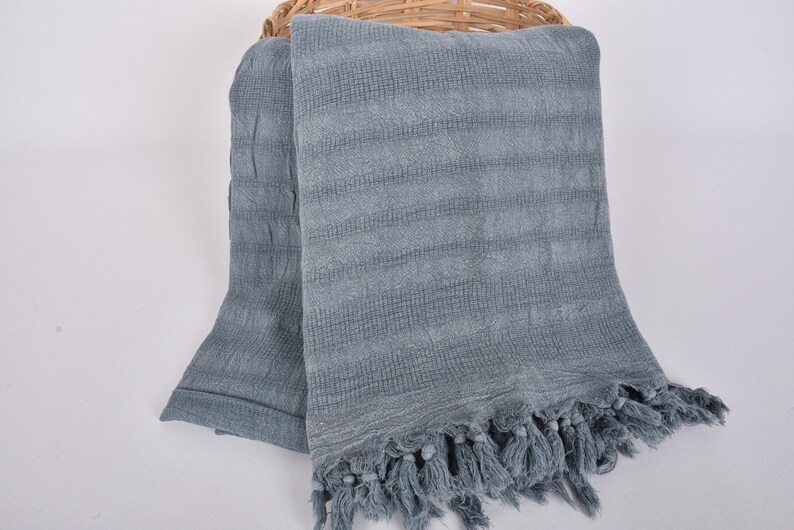 May include: A soft, blue, woven throw blanket with a fringe border. The blanket is folded and draped over a wicker basket.