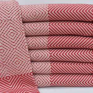 May include: A stack of six red and white patterned Turkish towels with fringe edges. The towels are folded and stacked on a white surface.