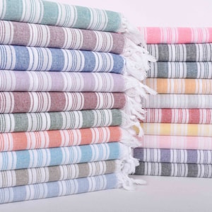 Puede incluir: Una pila de toallas turcas a rayas en varios colores, incluyendo verde petr&oacute;leo, azul marino y rosa brillante. Cada toalla tiene un borde con flecos y una etiqueta que indica el nombre del color. Las toallas est&aacute;n cuidadosamente dobladas y apiladas.