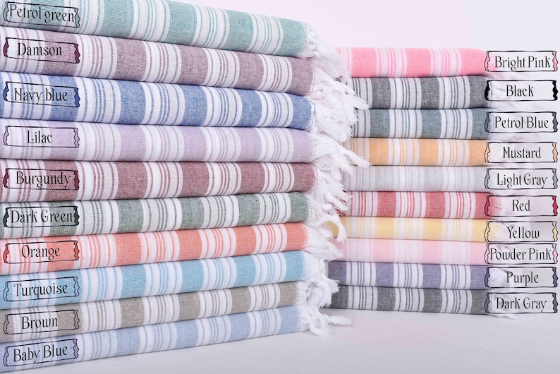 Puede incluir: Una pila de toallas turcas a rayas en varios colores, incluyendo verde petr&oacute;leo, azul marino y rosa brillante. Cada toalla tiene un borde con flecos y una etiqueta que indica el nombre del color. Las toallas est&aacute;n cuidadosamente dobladas y apiladas.