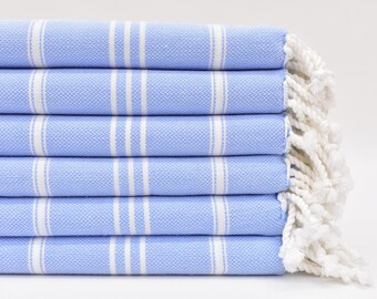 Può includere: Un mucchio di sei asciugamani di colore azzurro chiaro con strisce bianche e frange bianche. Gli asciugamani sono piegati e impilati su una superficie bianca.