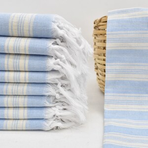 May include: A stack of light blue and white striped towels with white fringe. The towels are folded and stacked on a white surface.