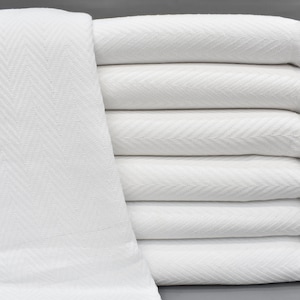 Può includere: Un mucchio di coperte bianche in cotone con un motivo a spina di pesce e bordi con frange.