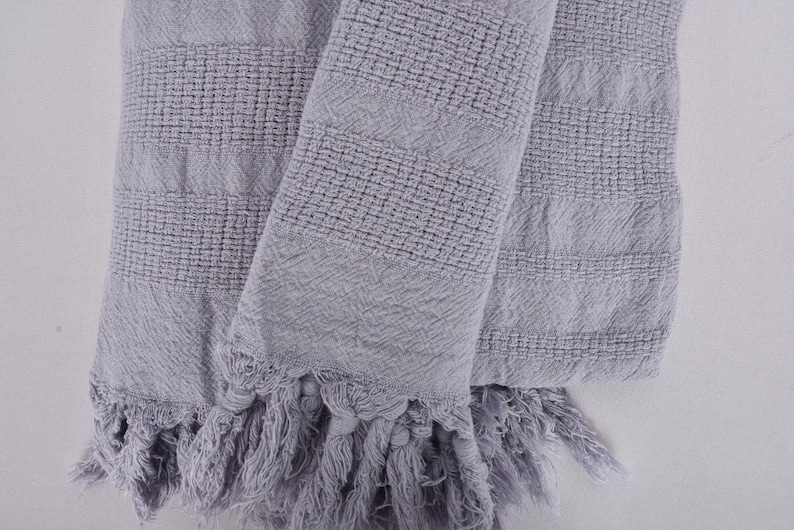 May include: A gray woven throw blanket with fringe on the edges. The blanket is folded in half and has a textured pattern.
