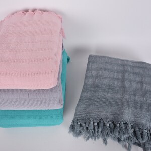 May include: A stack of three towels in pink, gray, and teal colors. The towels have a woven texture and fringe edges. A fourth towel in gray with fringe edges is lying flat next to the stack.