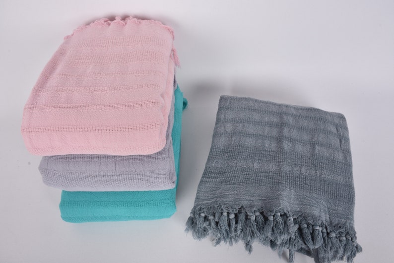 May include: A stack of three towels in pink, gray, and teal colors. The towels have a woven texture and fringe edges. A fourth towel in gray with fringe edges is lying flat next to the stack.