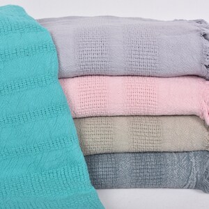 May include: A stack of five woven cotton throws with fringed edges. The throws are in shades of teal, light gray, beige, pink, and lavender.