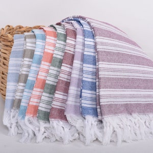 Puede incluir: Una colecci&oacute;n de toallas a rayas con flecos en varios colores, incluyendo azul, gris, naranja y morado, se exhiben junto a una cesta de mimbre. Las toallas tienen rayas horizontales blancas.